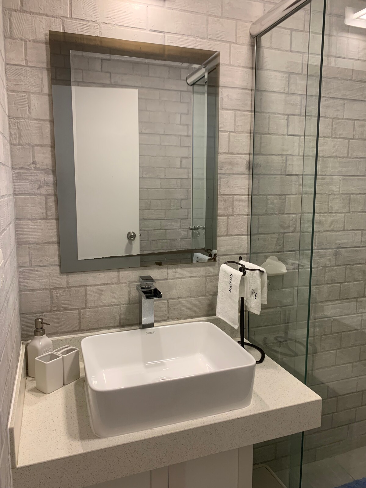 A modern bathroom features a sleek, square sink atop a light-colored countertop. A frameless glass shower enclosure is visible, alongside a neatly arranged set of toiletries and towels. A simple mirror reflects the space, complemented by textured wall tiles in neutral tones.