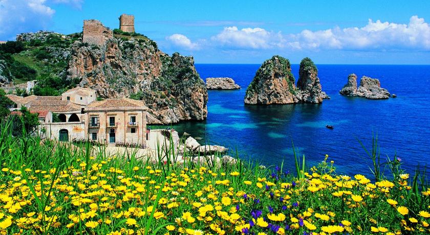A stunning coastal scene features rocky cliffs rising from a vibrant blue sea, accompanied by scattered islands. In the foreground, a colorful array of wildflowers adds a lively touch to the landscape. The sky is bright with scattered clouds, providing a serene backdrop.