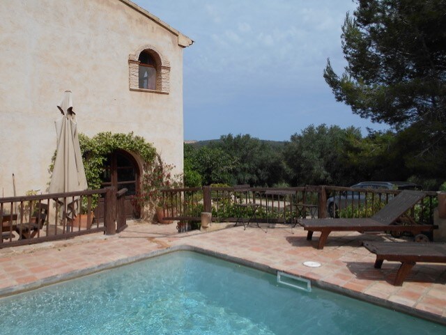 A rustic stone house is visible beside a clear swimming pool. A shaded area is provided by an umbrella, and comfortable lounge chairs are arranged along the poolside. Lush greenery and trees surround the outdoor space, creating a private and tranquil environment.