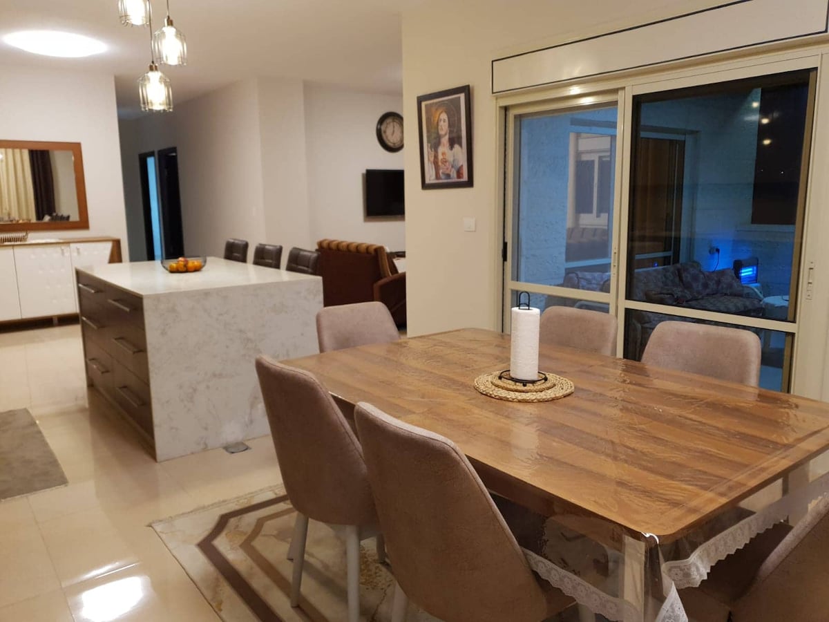 A dining area is equipped with a wooden table surrounded by soft chairs. A marble island is visible in the background, alongside a living area featuring a couch. Large glass doors lead to a balcony, allowing natural light to illuminate the space.