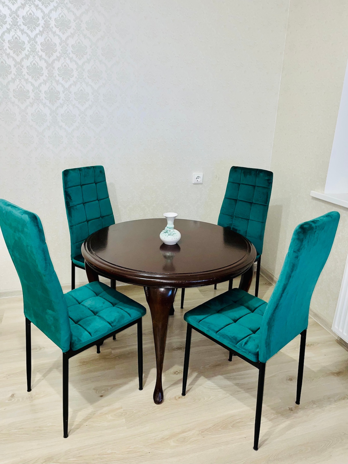 A round wooden dining table is surrounded by four upholstered chairs with a deep teal color. A small decorative vase sits at the center of the table. The background features light-colored, textured wallpaper, and the floor has a light wooden finish.