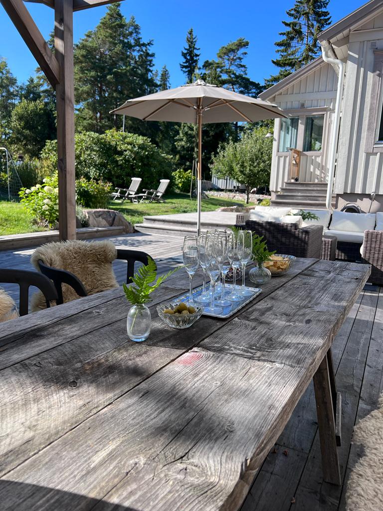 A rustic wooden table is set outdoors, adorned with glasses, a small vase with greenery, and a platter of olives and snacks. An umbrella provides shade, with comfortable seating visible nearby and a well-maintained lawn in the background under clear blue skies.