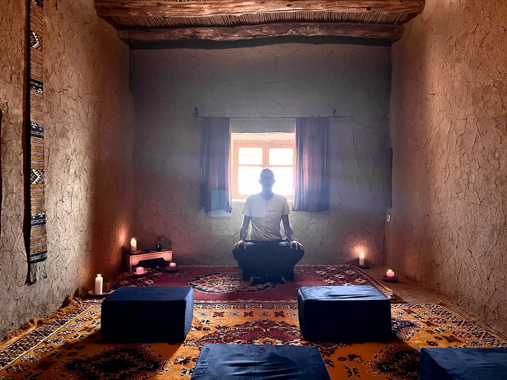 A serene interior space features an earthen wall and wooden ceiling beams. A small window allows soft light to filter in, illuminating the area. Cushions are arranged on a vibrant patterned rug, complemented by dim candlelight for a calming atmosphere.