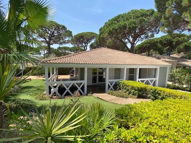 A charming chalet displays a sloped roof covered with terracotta tiles, surrounded by lush greenery and palm trees. Multiple pathways lead to a welcoming veranda, with a landscaped garden featuring vibrant shrubs and neatly trimmed grass.