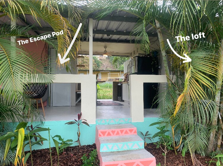 The entrance to the property is showcased, revealing two separate units labeled 'The Escape Pod' and 'The Loft'. Lush tropical plants frame the pathway, while a modern kitchen is visible through the open space. A light concrete surface connects the units, enhancing the inviting atmosphere.