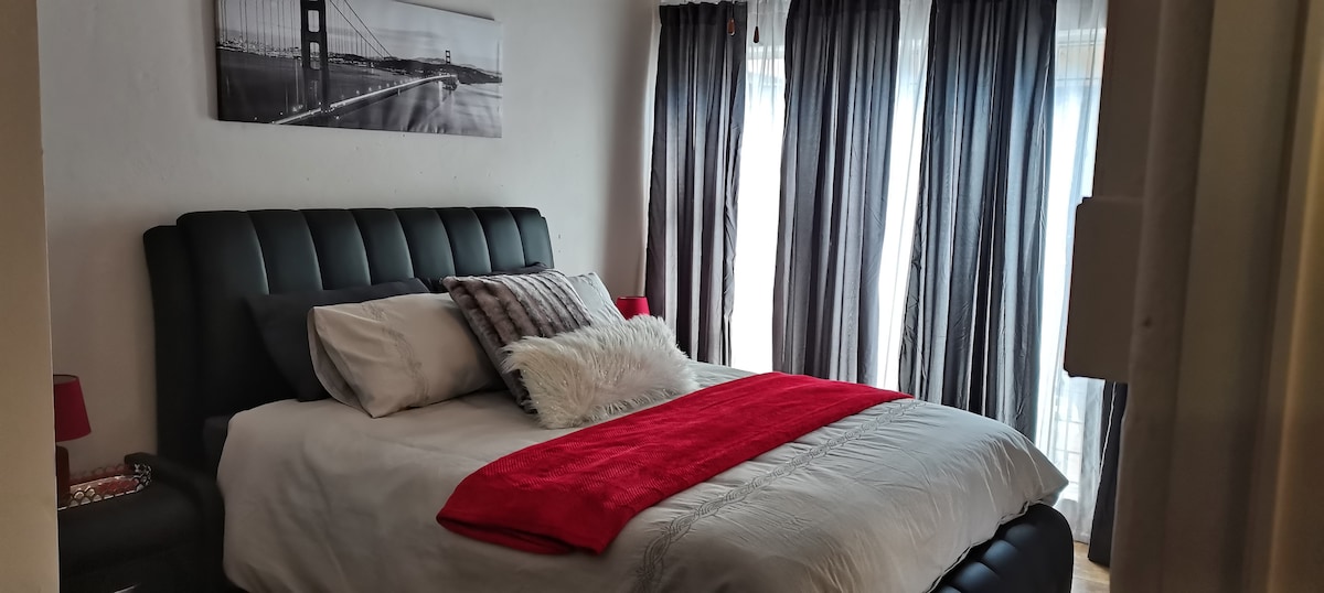 A spacious bedroom features a large bed dressed in neutral linens, accented with a vibrant red throw. Dark curtains frame the windows, allowing soft light to filter in. A modern black headboard complements the overall design, while a cityscape photograph adorns the wall above.