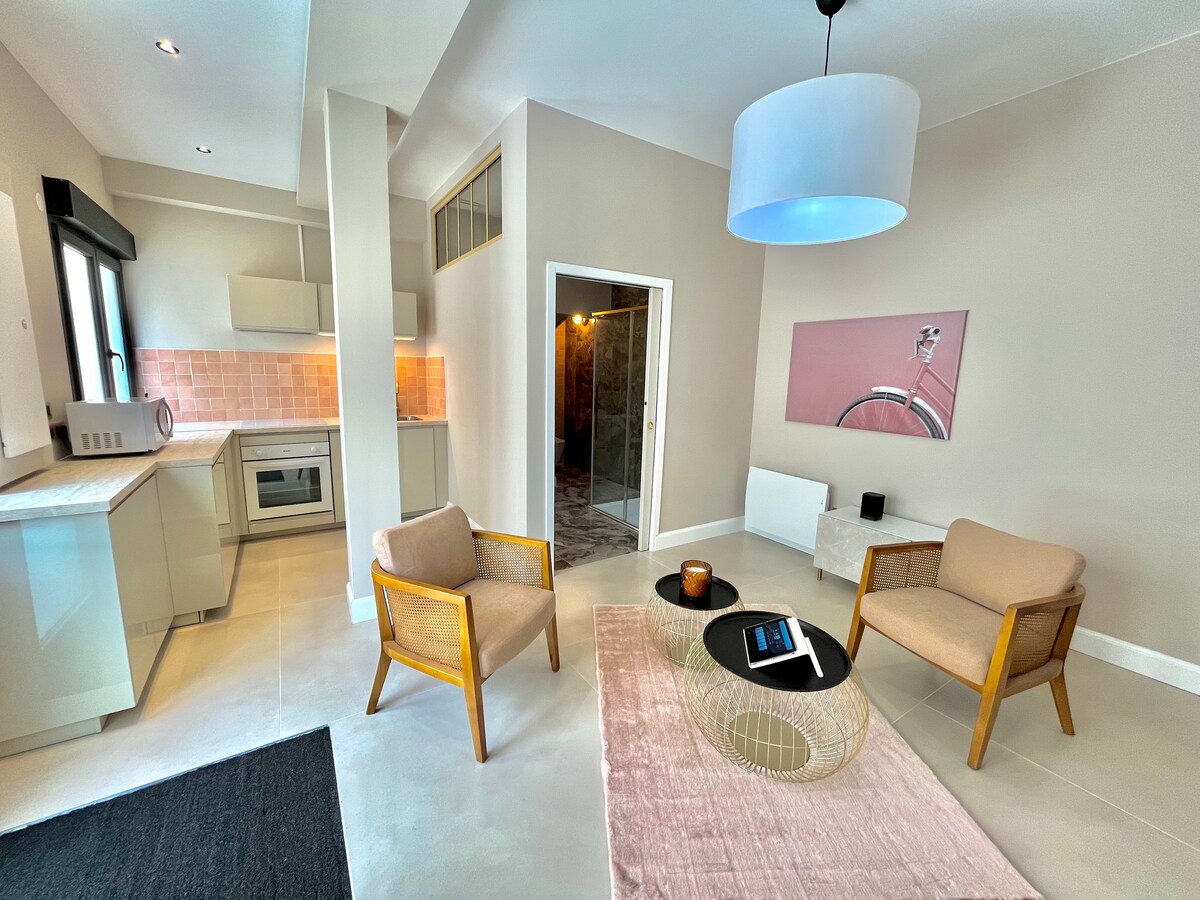 A modern living area features a light-colored decor with two wooden chairs and a coffee table at the center. A sleek kitchenette is visible in the background, accented by warm lighting. A doorway leads to the bathroom area, and a pink art piece hangs on the wall.