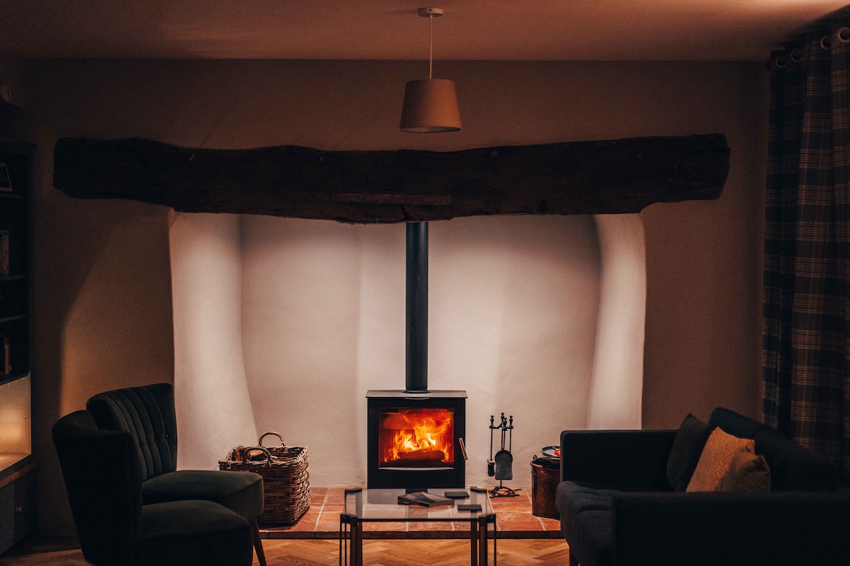 A wood burning stove is centered against a smooth, light-colored wall, framed by rustic wooden beams. The cozy seating area features two green armchairs and a coffee table, with a warm glow from the fire creating a welcoming atmosphere.