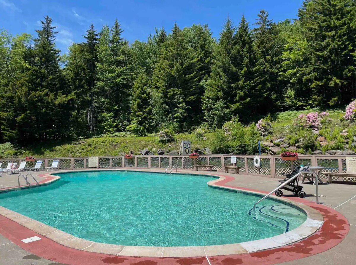 An inviting outdoor swimming pool is surrounded by a textured concrete deck, featuring a gentle slope for entering the water. Lush greenery and vibrant flowers create a natural backdrop, while lounge chairs are positioned at the pool’s edge for relaxation.