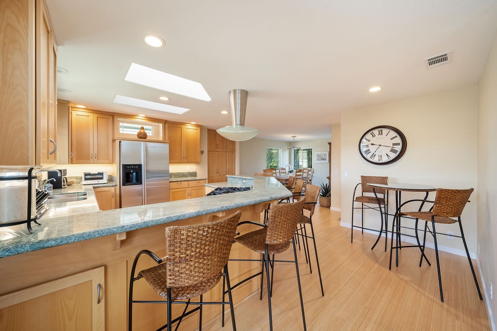 A modern kitchen is equipped with wood cabinetry and a large granite island with seating for four. Stainless steel appliances are complemented by a built-in microwave and spacious countertops. Natural light filters through multiple skylights, enhancing the open layout that connects to the dining area.