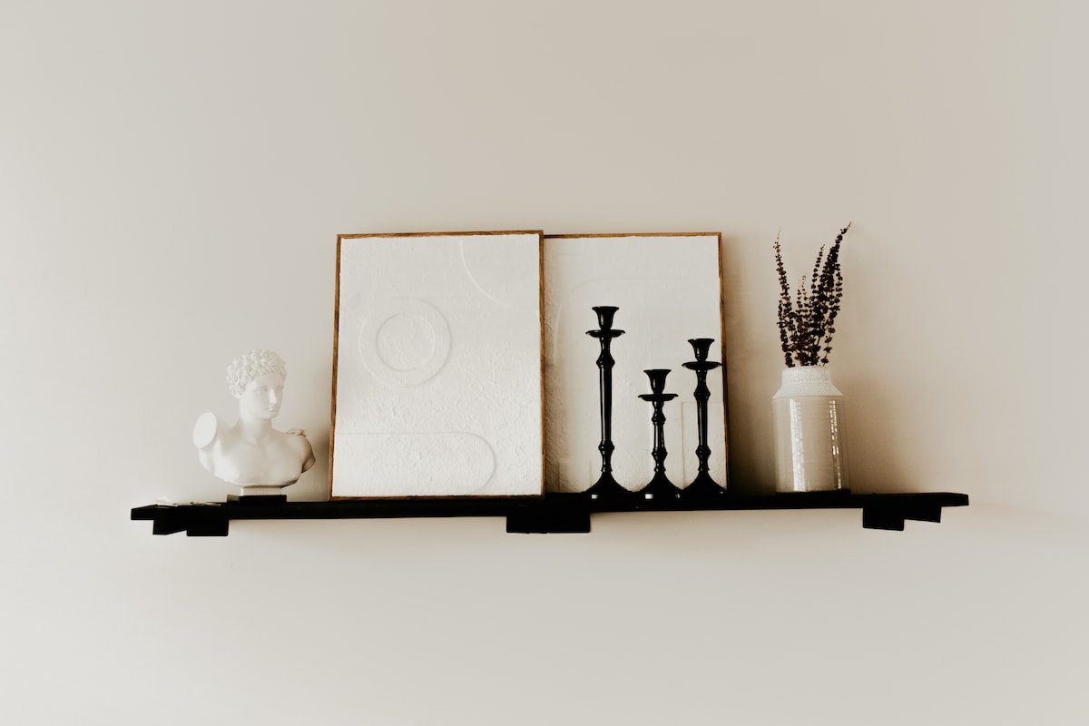 A wall-mounted shelf displays a decorative white teapot, two framed artworks, and a slender vase filled with dried flowers. Three black candle holders are positioned on the shelf, contributing to the minimalist aesthetic of the space.