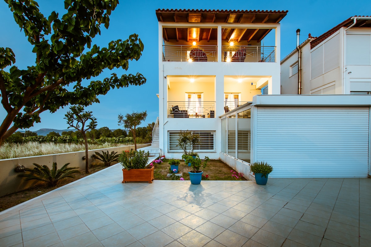 The exterior of Erato Suite is showcased, highlighting a two-level structure with spacious balconies and large windows. A tiled patio is seen in the foreground, flanked by potted plants. Lush greenery and trees add to the serene ambiance of the property.