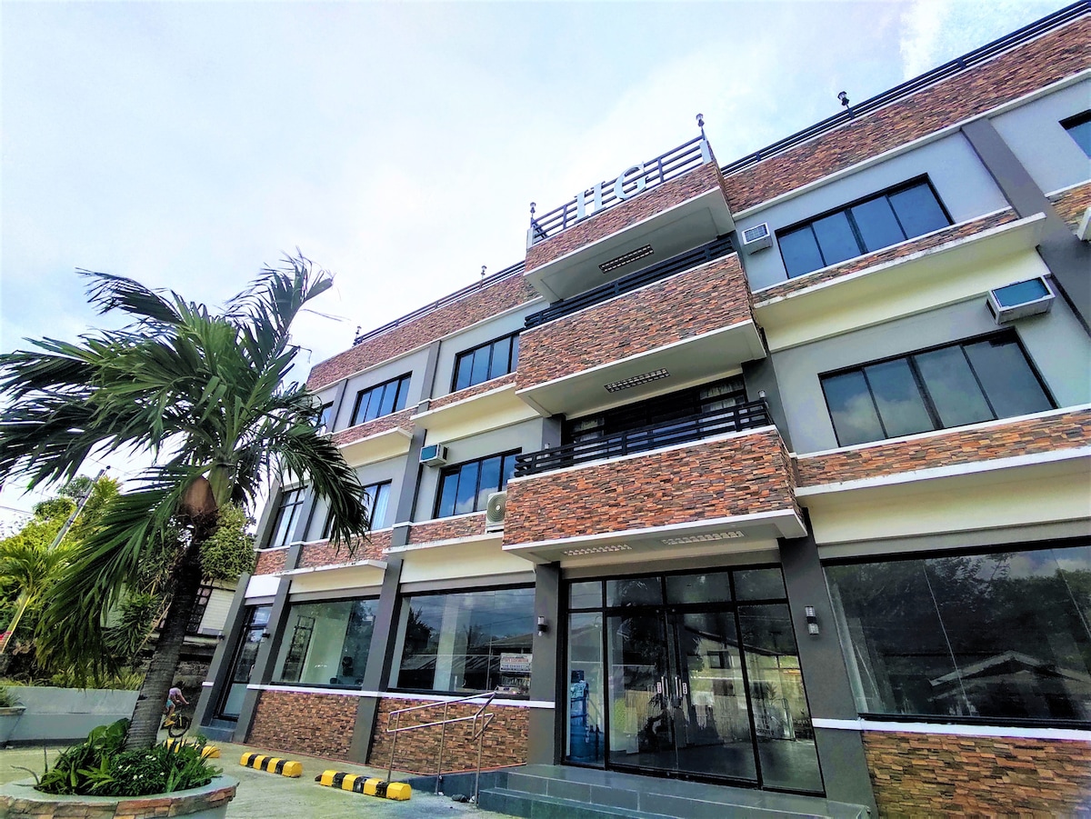 An exterior view of a modern multi-story building features a combination of stone and glass elements. Large windows provide ample natural light, while a palm tree adds a touch of greenery to the entrance area. The building showcases a welcoming atmosphere.