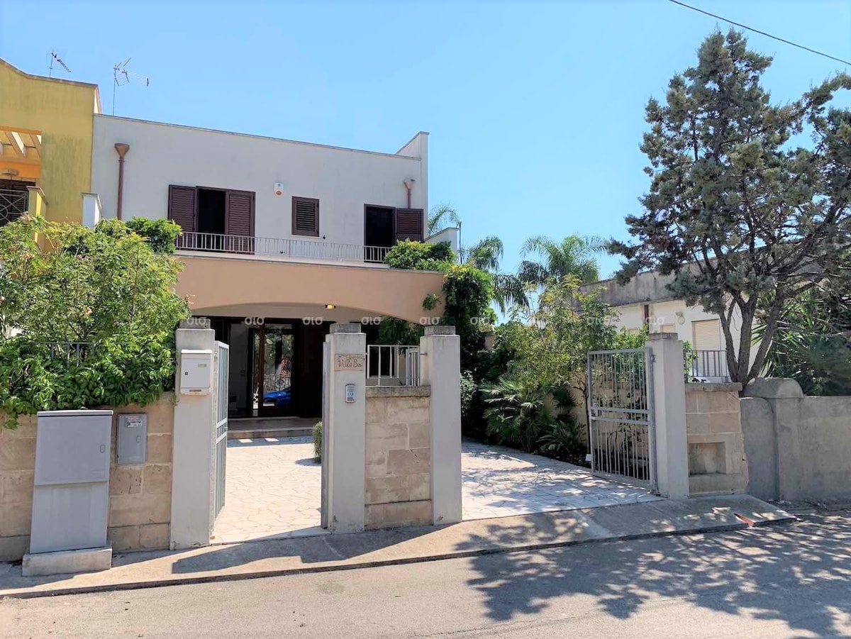 A welcoming entrance to the property is framed by greenery and decorative stones. The two-story building features a wide archway and classic shutters. A pathway leads towards the gate, with tropical plants enhancing the outdoor space.