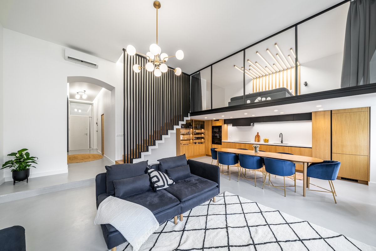 A modern living space features an open layout with a spacious sofa, a light rug, and a dining area with blue chairs. Stairs lead to a second level, while a well-equipped kitchen with wooden cabinetry is visible in the background. Natural light fills the area.