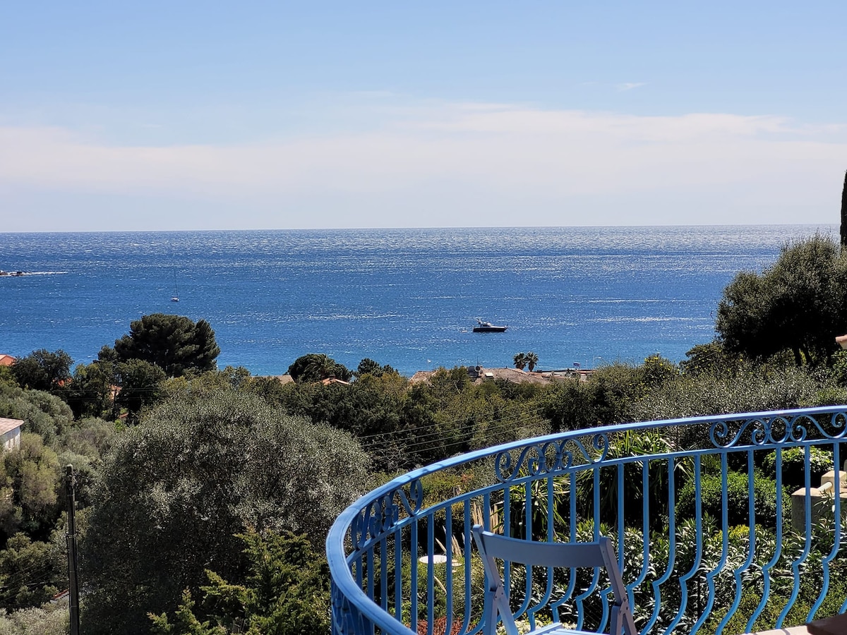 A view overlooking the sea from a blue wrought-iron balcony, where vibrant greenery is present. The calm water reflects sunlight, and a boat can be seen in the distance, enhancing the serene coastal landscape.