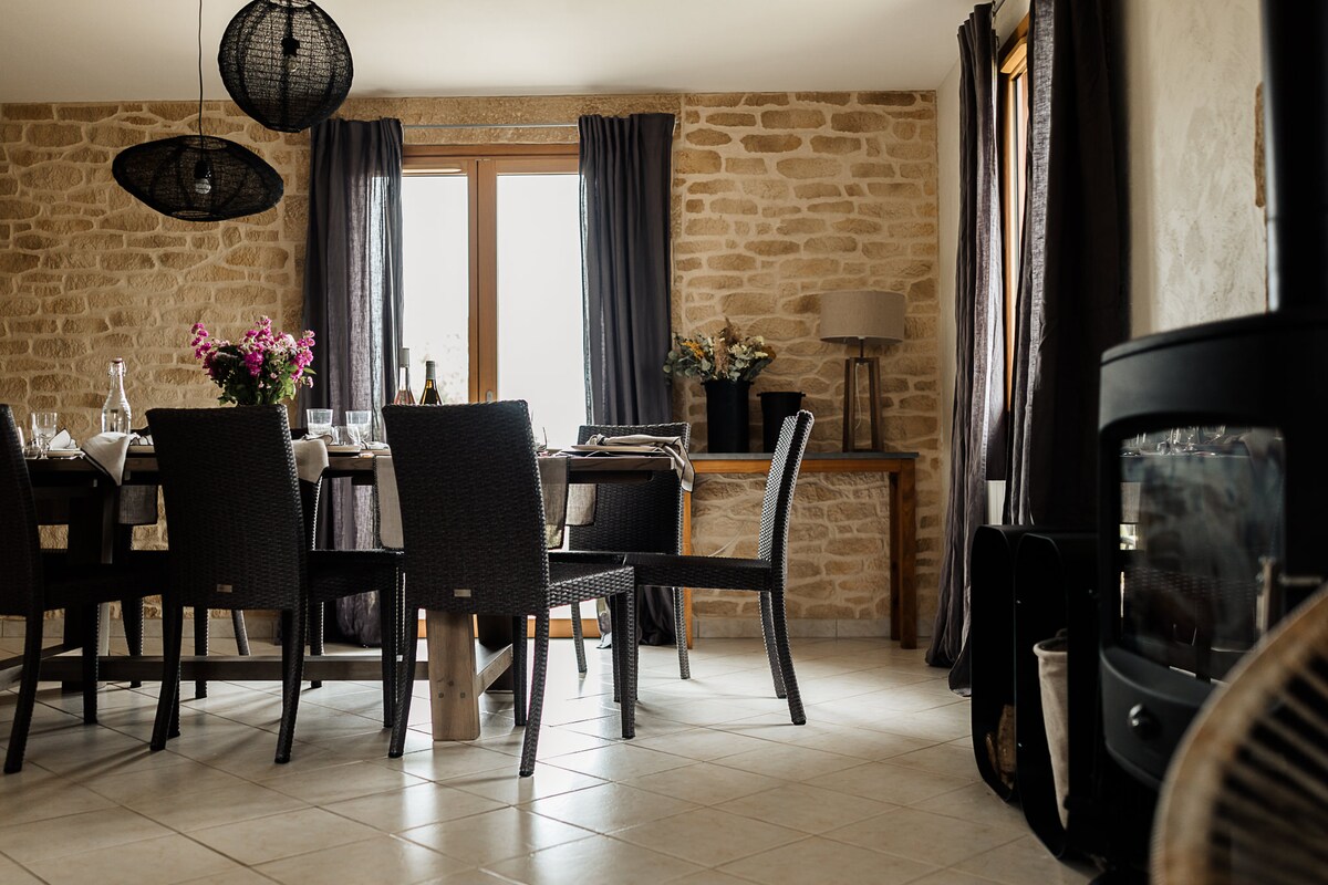 A spacious dining area features a large table surrounded by dark wicker chairs. Natural light enters through a window, which is framed by dark curtains. A wood stove is positioned nearby, with decorative elements, including vases of flowers, adding a welcoming touch.