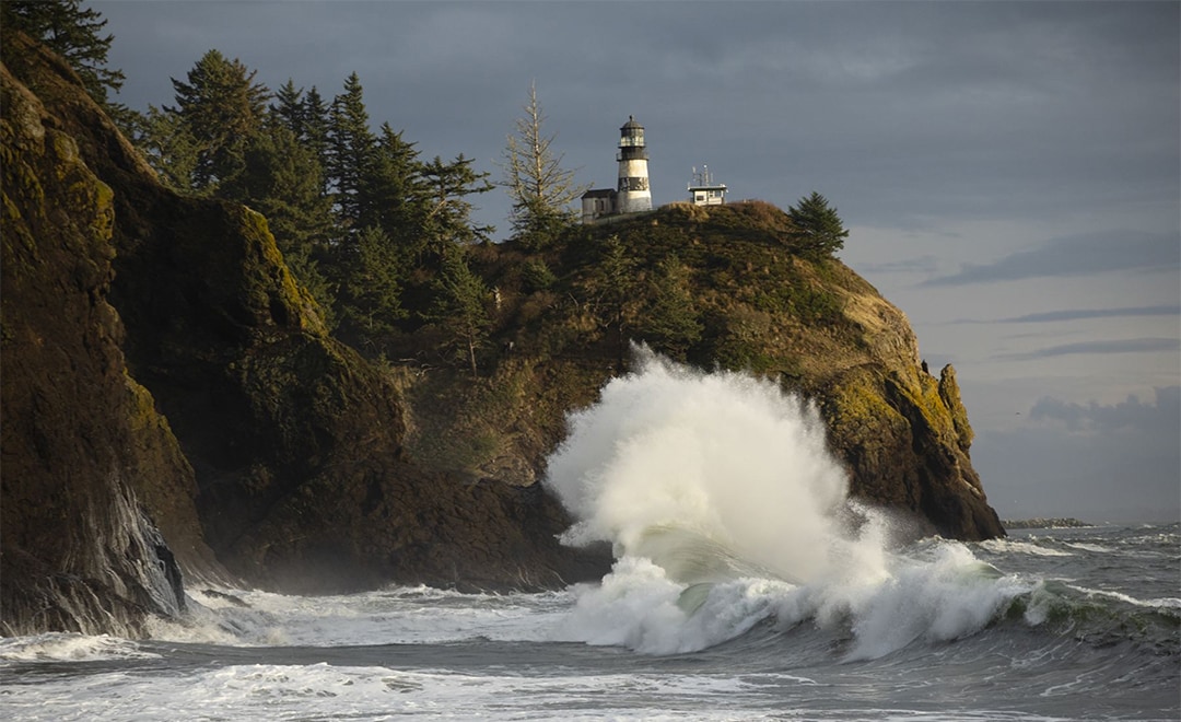 A lighthouse atop a rocky outcrop overlooks crashing waves. Tall trees surround the cliff, while the ocean displays white-capped waves splashing against the shore. The sky is overcast, enhancing the rugged coastal scenery.