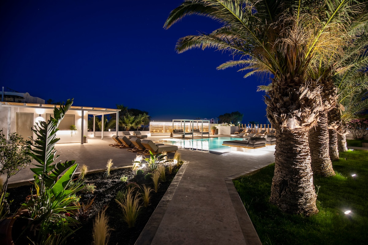 An outdoor pool area is surrounded by palm trees, with lounge chairs arranged for relaxation. Soft lighting illuminates the space, creating a serene atmosphere. Nearby, a bar area is visible, enhancing the social setting of the elegant poolside retreat.