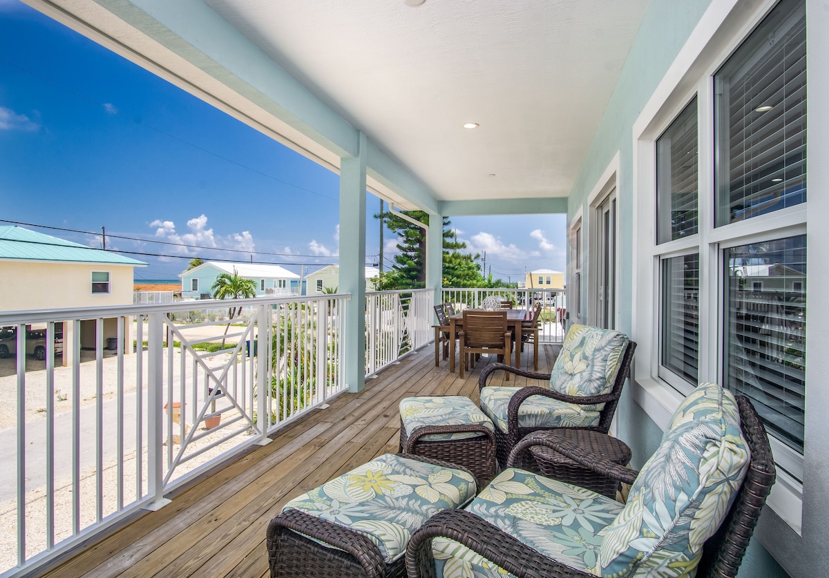 A spacious balcony showcases comfortable wicker chairs with tropical-patterned cushions, ideal for relaxation. A wooden dining table accompanies the seating area, complemented by scenic views of the surrounding neighborhood and blue sky. The space is illuminated by natural light, enhancing the outdoor atmosphere.