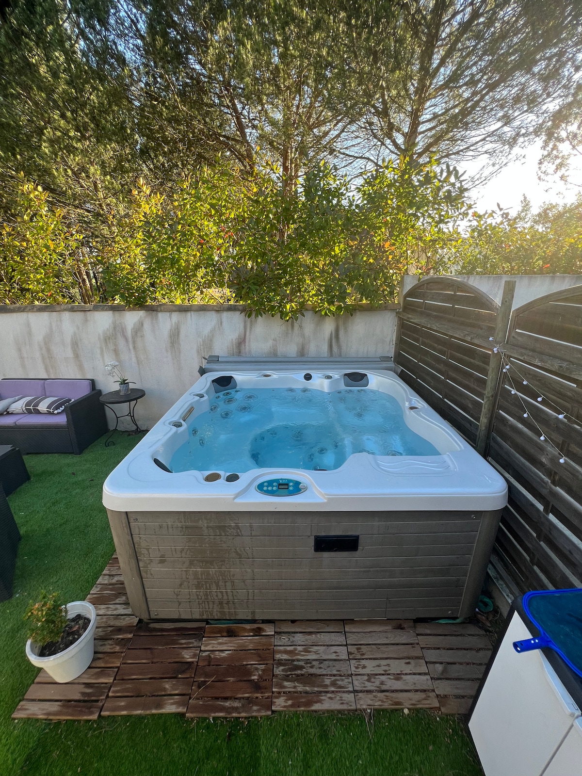 A spacious jacuzzi is surrounded by wooden decking and greenery, creating a private outdoor retreat. Nearby, a small plant sits in a white pot, adding a touch of nature. The area is framed by wooden panels, enhancing the sense of seclusion.