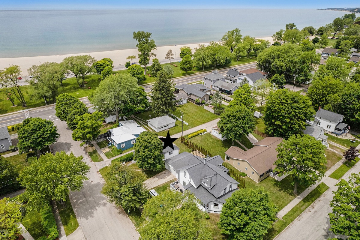 An aerial view showcases a serene neighborhood with green trees lining the streets. In the background, the sandy beach and blue water are visible, illustrating the close proximity to the shore. The layout of nearby homes and the avenue is clearly defined.