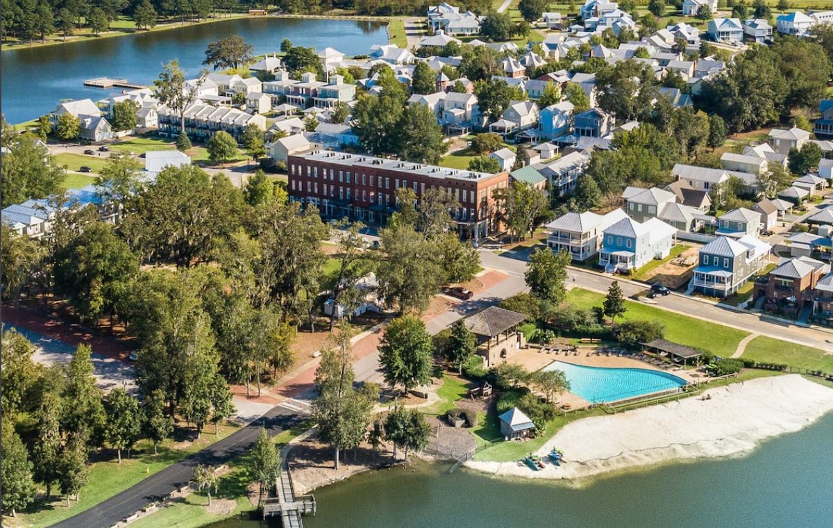 An aerial view showcases the community, featuring multiple residential homes, lush green areas, and three lakes. A swimming pool is visible near the water's edge, surrounded by trees, with a sandy beach area leading to the lake.