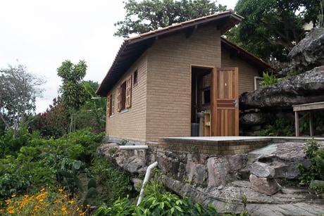 A charming stone and brick chalet is nestled among lush greenery and flowering plants. A wooden door leads to the interior, and large windows allow natural light to fill the space. The surrounding rocks and vegetation create a serene outdoor environment.