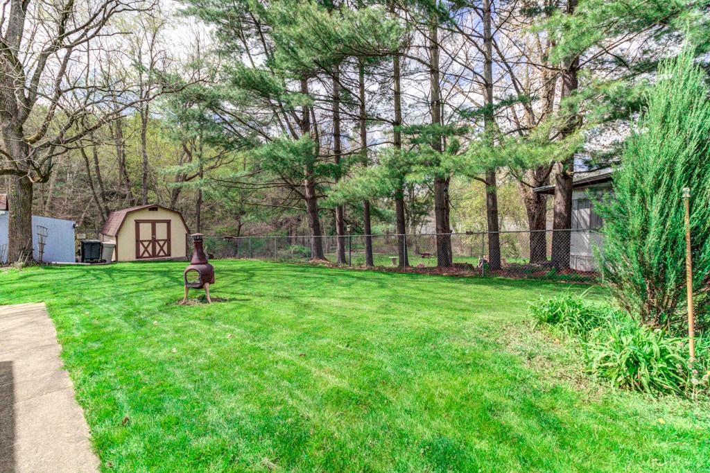 A large fenced yard is displayed, featuring lush green grass and tall trees along the perimeter. A storage shed with a charming door is visible, and a fire pit is centered in the yard, providing a space for gatherings. The area remains quiet and spacious.