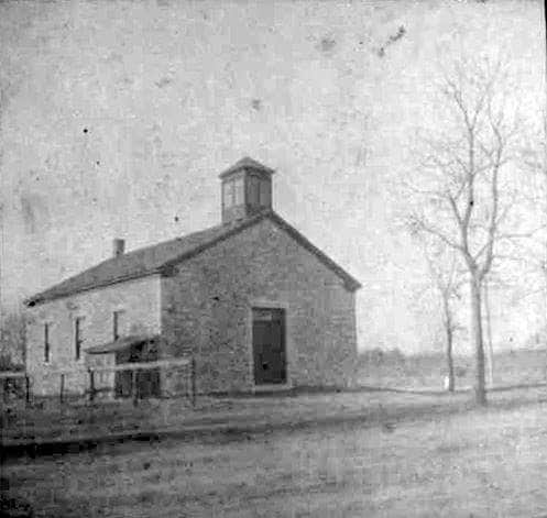 The historic building is shown in a monochromatic view, presenting its stone exterior and simple architecture. A single tree stands nearby, with the road visible in the foreground. The structure features a gabled roof and a small cupola at the top.