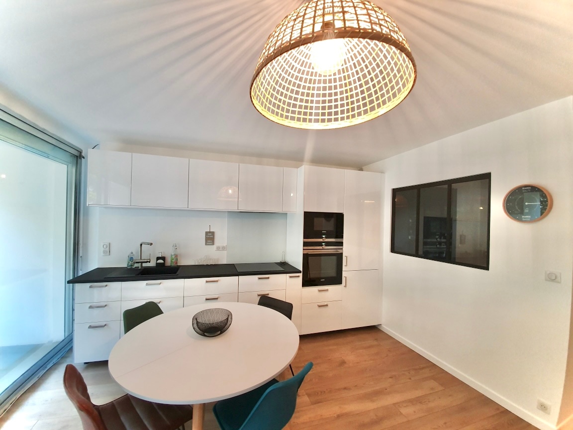 A modern kitchen is presented with sleek white cabinetry and a black countertop. A round dining table is set in the center, surrounded by colorful chairs. A large window floods the space with natural light, while a distinctive pendant light adds visual interest overhead.