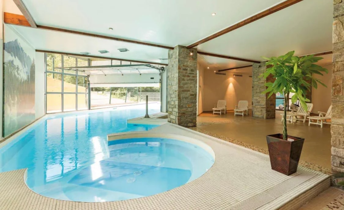 An indoor swimming area features a semi-covered heated pool with a whirlpool section. A large glass wall provides views of the surrounding landscape. Several lounge chairs are positioned nearby, and a potted plant adds a touch of greenery to the space.