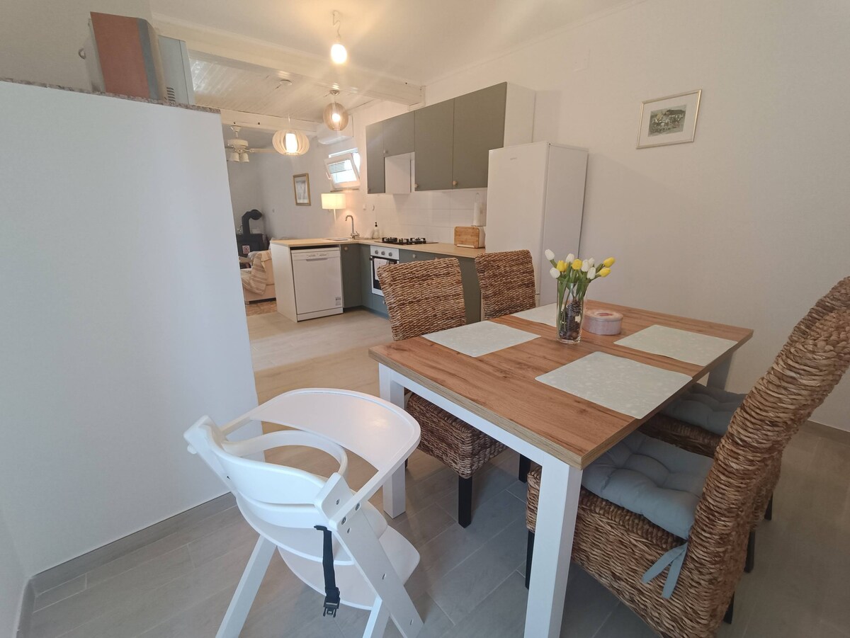 The open-plan kitchen and dining area features a wooden table set with light accessories. Four woven chairs surround the table, and a high chair is positioned nearby. Kitchen cabinets in soft colors are complemented by modern appliances, creating a functional yet inviting space.