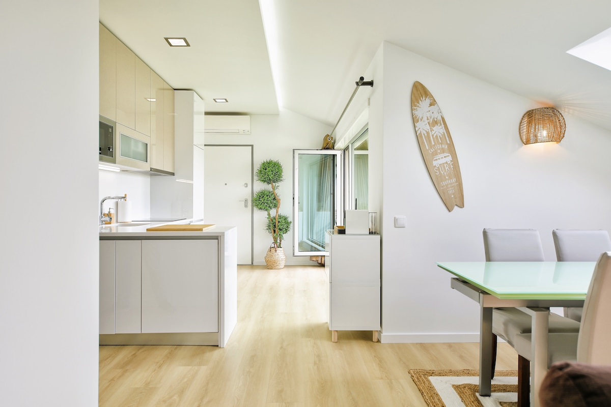 A modern kitchen and dining area are showcased, featuring sleek cabinetry and a glass-top dining table. Natural light fills the space through large windows, while decorative greenery adds a touch of freshness. A surfboard wall decoration enhances the coastal theme.