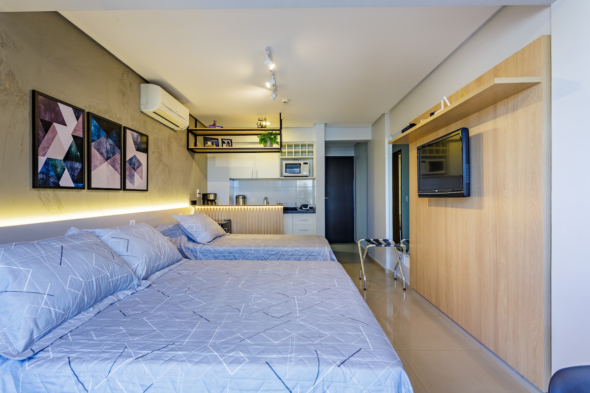The spacious room features two comfortable beds adorned with gray patterned bedding. A kitchenette is visible in the background, complete with modern appliances and a microwave. A flat-screen TV is mounted on the wooden accent wall, while soft lighting highlights the attractive décor.