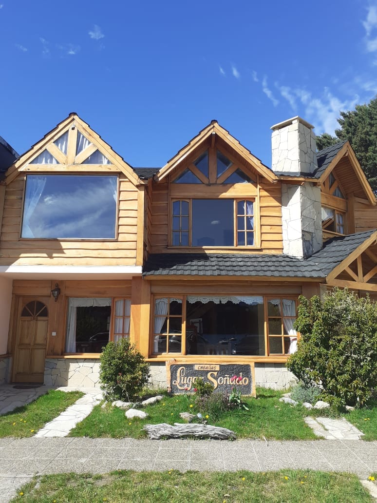 The exterior of a wooden cabin features a charming facade with peaked roofs and large windows. A stone chimney rises above, surrounded by green shrubs and a well-maintained garden. A sign reading 'Cabañas Lugar Soñado' is visible in front, complemented by a clear blue sky.