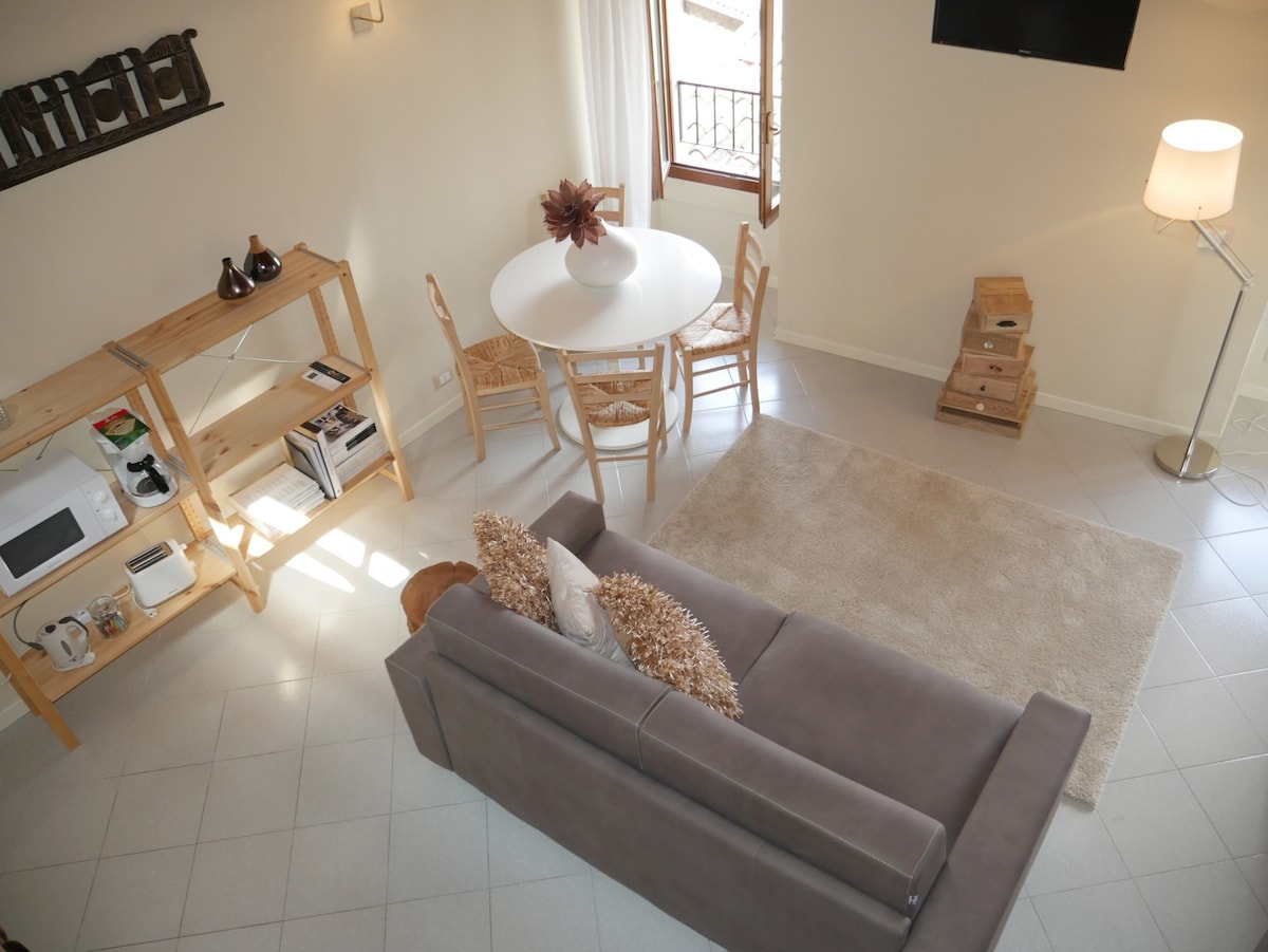 A well-lit living area is characterized by a soft fabric sofa facing a circular dining table surrounded by four chairs. A light-colored area rug is placed under the seating, while a floor lamp and wooden shelving units provide additional functionality and warmth.