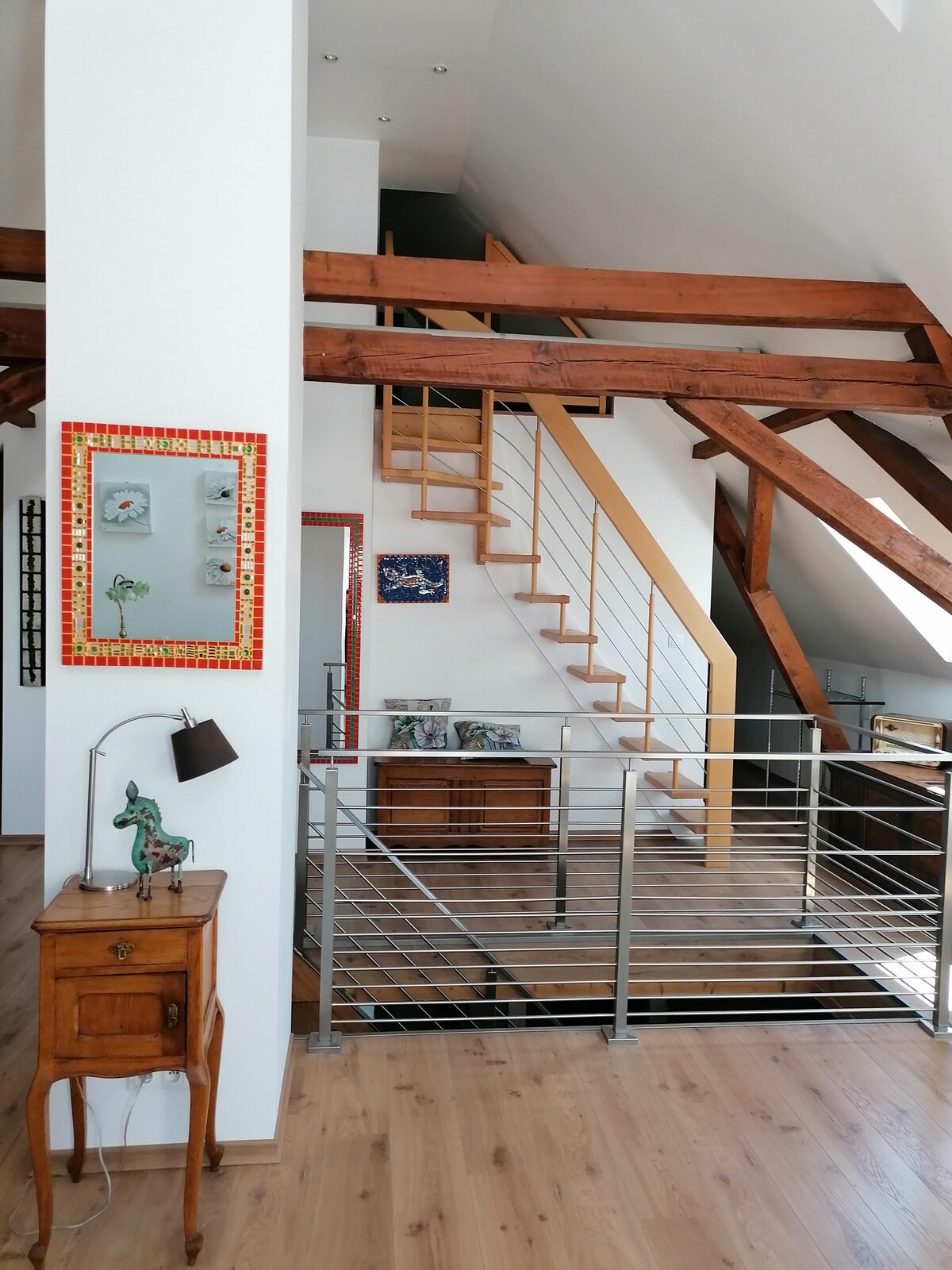 An open stairway features wooden steps leading to an upper level, framed by a sleek metal railing. Natural light filters through skylights, illuminating the wooden beams above. A small table with a decorative lamp stands beside a mirror, adding character to the airy space.