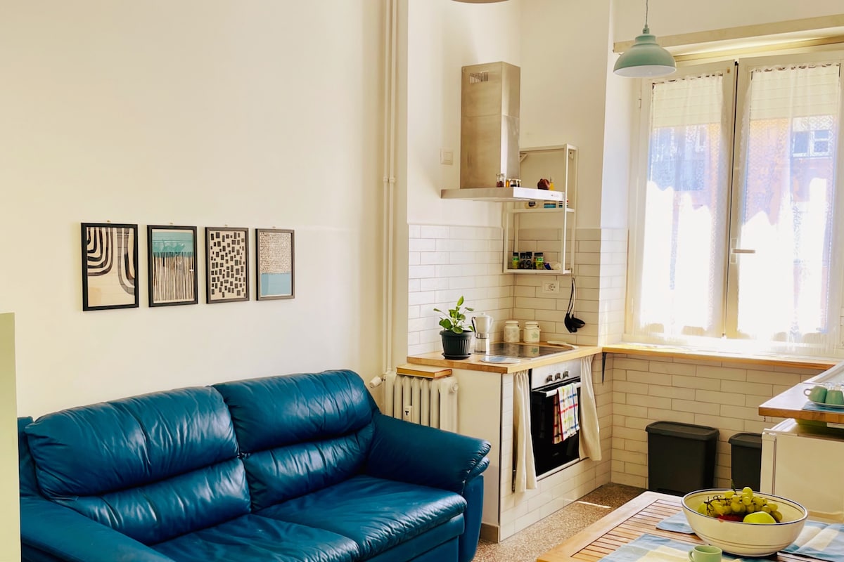 A cozy living space is visible with a blue sofa facing a wall adorned with framed artworks. A kitchen area features a stove, sink, and shelving above a wooden countertop. Natural light fills the room through large windows, enhancing the bright ambiance.