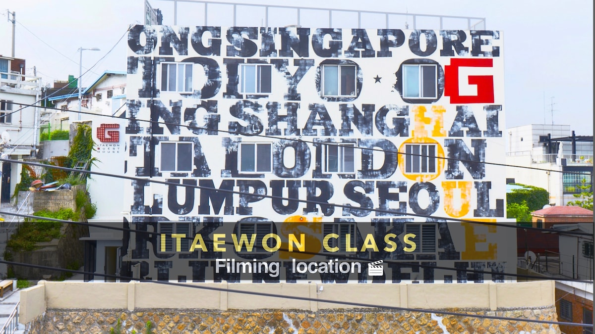 A large mural displaying the names of various cities the guesthouse is associated with, prominently featuring 'SEOUL' and 'G' in bold lettering. The mural serves as a notable backdrop and is part of the unique architecture surrounding the guesthouse.