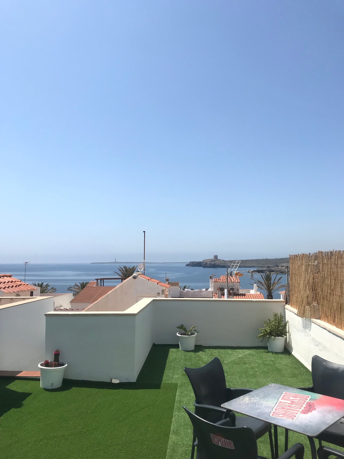 A spacious terrace features artificial grass and potted plants, offering a panoramic view of the sea and surrounding rooftops. The calming blue sky sets a serene backdrop, while distant coastal structures can be seen on the horizon.