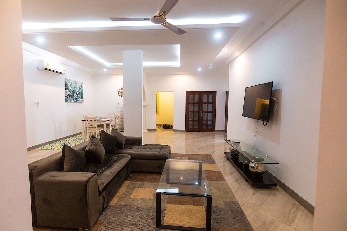 A spacious living area is presented with a sectional sofa in a neutral tone, complemented by a glass coffee table. Soft lighting highlights the sleek flat-screen TV mounted on the wall, while the open layout connects to a dining space and kitchen area.