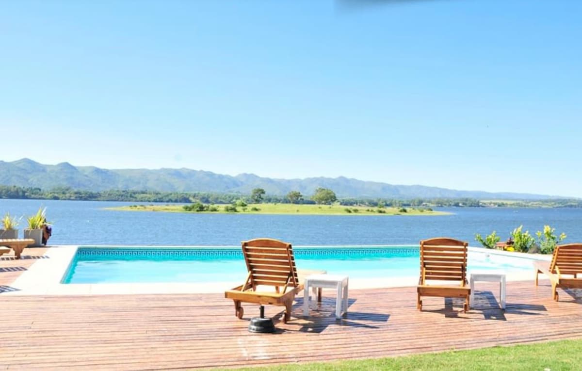 A serene outdoor space features a pool bordered by wooden decking. Sun loungers are positioned for relaxation, overlooking the peaceful lake. Lush greenery and distant mountains can be seen against a clear blue sky, creating a tranquil atmosphere.