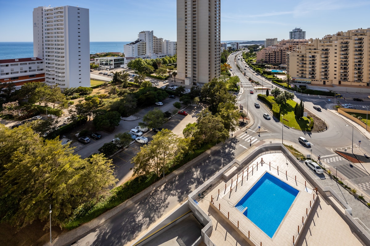 A panoramic view captures the pool area surrounded by neatly arranged sun loungers. The ocean is visible in the distance, framed by tall buildings. A landscaped area with trees and open parking is seen below, illustrating the neighborhood's accessibility and inviting environment.