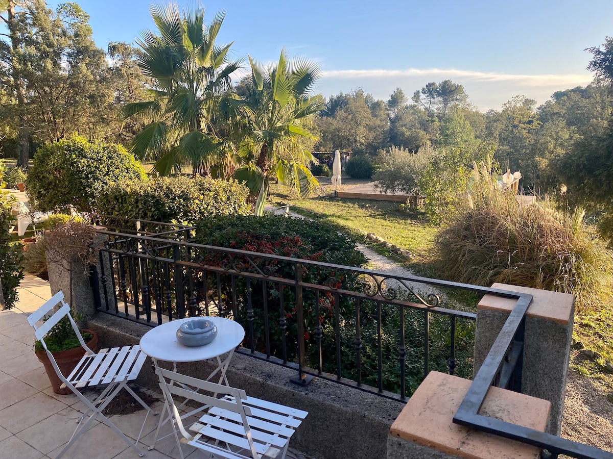 A private terrace features a round table with a bowl and two white chairs, surrounded by lush greenery and trees. The view showcases a spacious garden with various plants in the background, providing a calm and serene outdoor atmosphere.