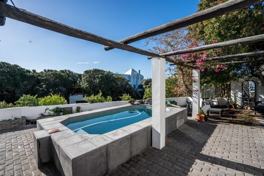 An outdoor pool area features a rectangular pool with a concrete surround, shaded by wooden beams. Lush greenery and flowering plants create a natural backdrop, enhancing the spacious patio made of brick. Comfortable seating arrangements are visible nearby, inviting relaxation in a tranquil setting.