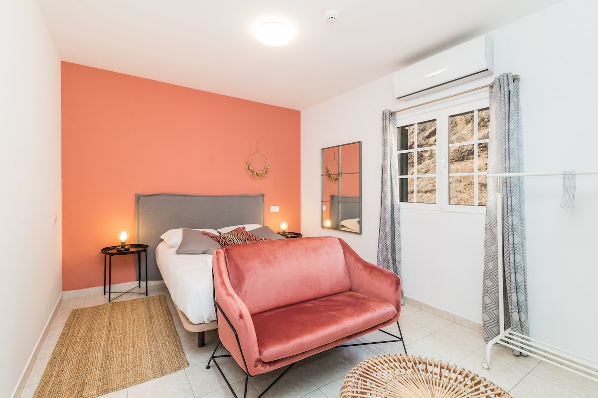 A bright private suite features a spacious bed with neutral linens, complemented by a coral accent wall. A plush pink sofa invites relaxation, while natural light enters through a window adorned with patterned curtains. An air conditioning unit and a decorative mirror enhance the room's comfort.