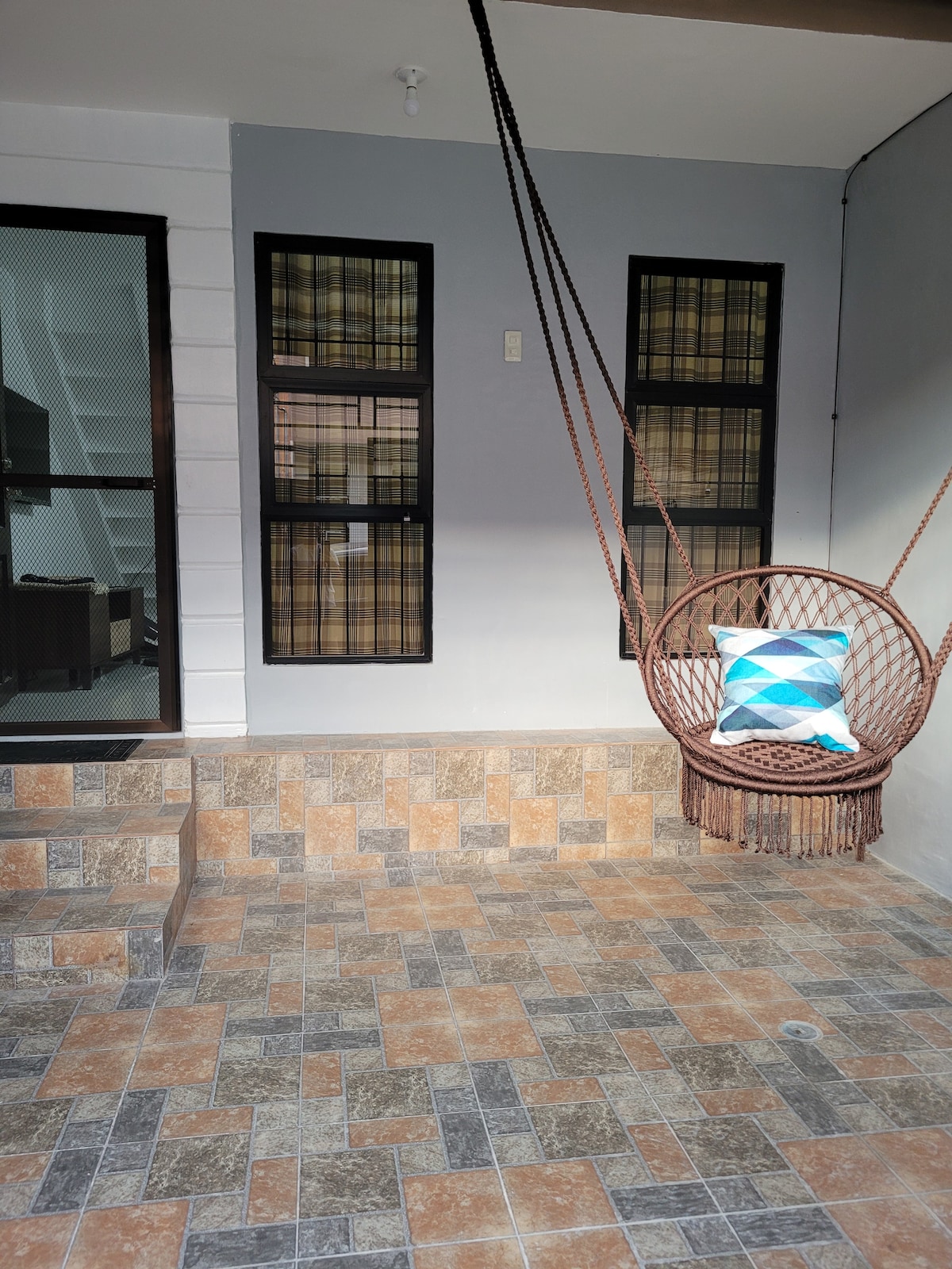 A cozy outdoor area features a decorative hanging chair with a geometric-patterned pillow. The stone-tiled floor is arranged in a checkerboard pattern, while large windows with plaid curtains are visible beside the front door, allowing natural light to enhance the space.