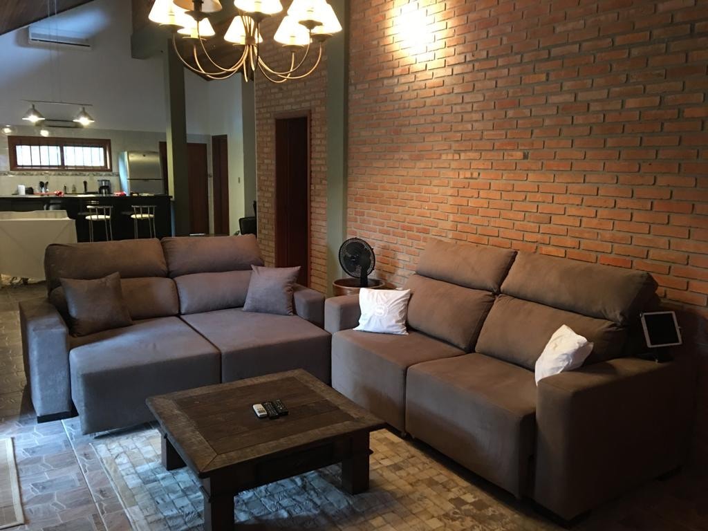 A comfortable seating area features two sectional sofas with soft, neutral fabric. A wooden coffee table is positioned in front, complemented by decorative pillows. A chandelier with multiple bulbs illuminates the warm brick wall, enhancing the cozy atmosphere. The kitchen can be partially seen in the background.