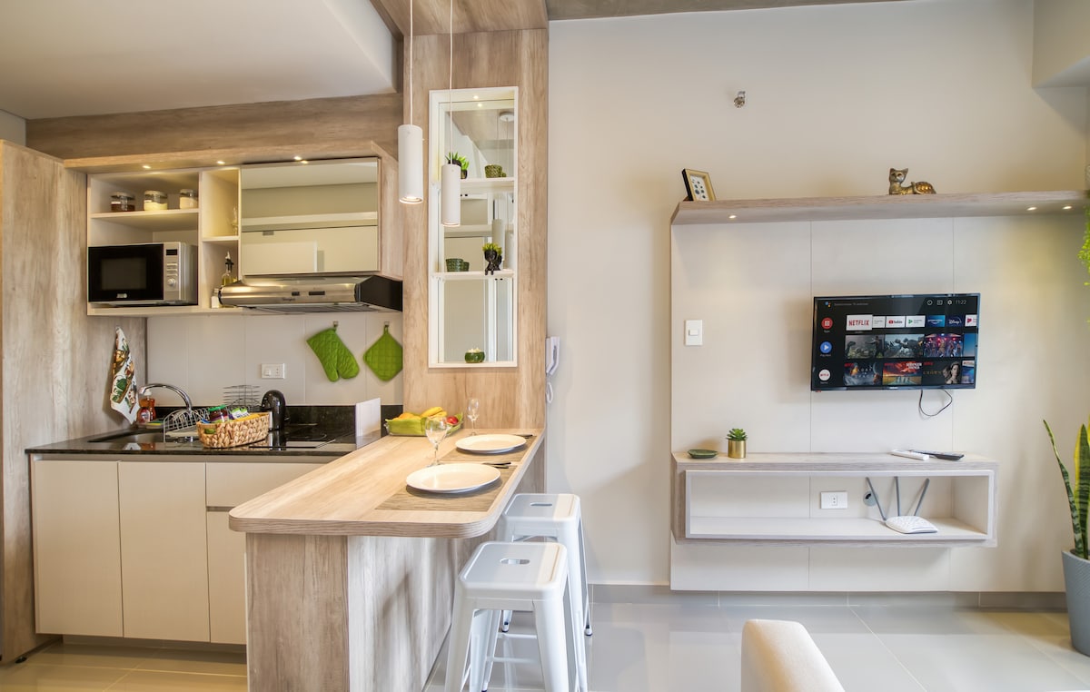 A modern kitchen space features light wood cabinetry and a streamlined design. The kitchen island with bar seating is paired with a dining setup. Appliances include a microwave and stove, while a wall-mounted TV is positioned nearby, complementing the inviting atmosphere.